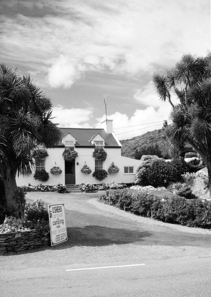 Zwart-wit foto van een Iers huisje in de zon voor een heuvel en een bewolkte lucht. Aan de weg staat een bord: 'O'SHEAS Camping and Caravaning Parc, Free Showers'.